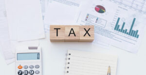Mesa de trabajo con documentos financieros, gráficos y una calculadora, junto a bloques de madera que forman la palabra “TAX” y un cuaderno abierto.