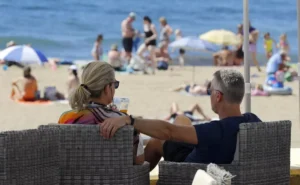 Pareja sentada en una terraza junto a la playa, observando el mar y la gente en la arena, en un ambiente relajado y soleado.