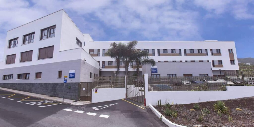 Edificio moderno de varias plantas, probablemente una residencia o centro sociosanitario, con fachada blanca, ventanas alineadas, palmeras en la entrada y acceso vallado desde la calle.
