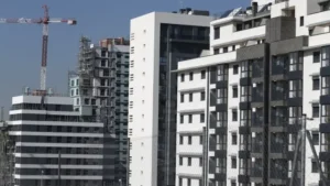 Vista de varios edificios residenciales modernos de varios pisos, con fachadas blancas y grises, junto a una grúa de construcción y estructuras en obra al fondo, bajo un cielo despejado.