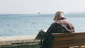Hombre mayor sentado en un banco junto al mar, leyendo un periódico, con el horizonte y el agua tranquila al fondo.