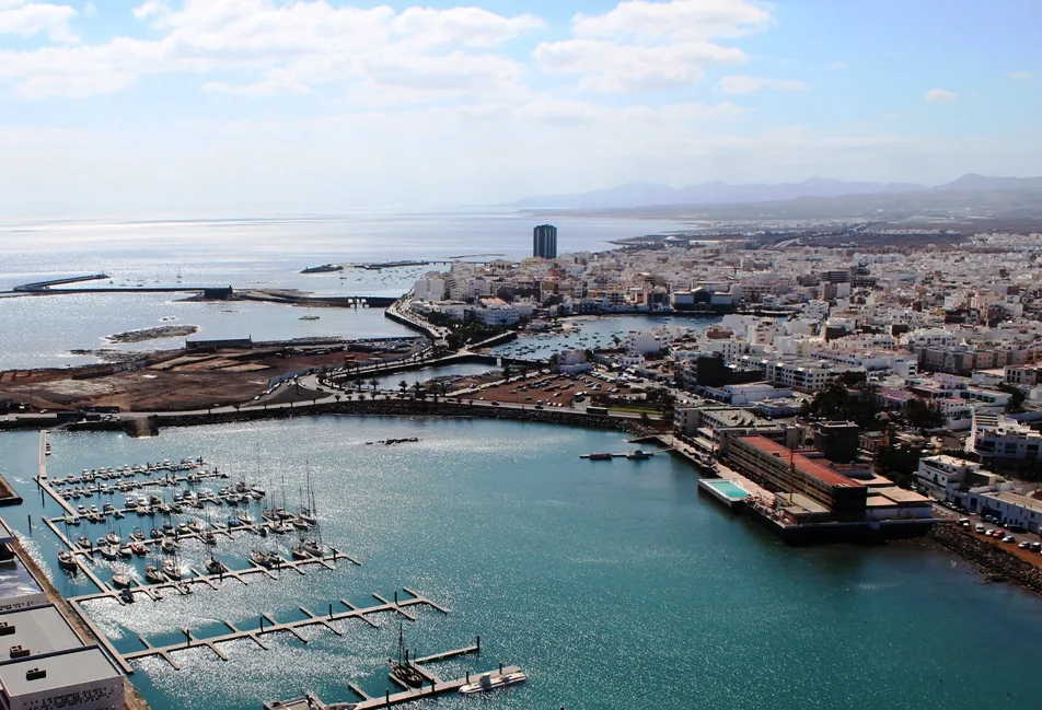 "Vista panorámica de una ciudad portuaria con marina, edificios blancos, costa atlántica y montañas al fondo bajo cielo azul."
