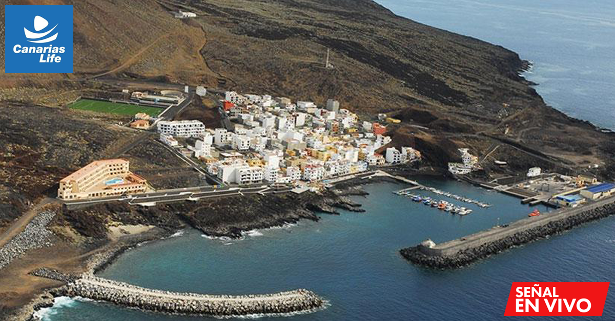 Vista aérea de un pueblo costero en las Islas Canarias con casas blancas agrupadas junto a un puerto deportivo protegido por rompeolas. El paisaje muestra terreno volcánico rocoso y montañas áridas. Se observa un campo de fútbol, edificios residenciales y embarcaciones en el muelle.