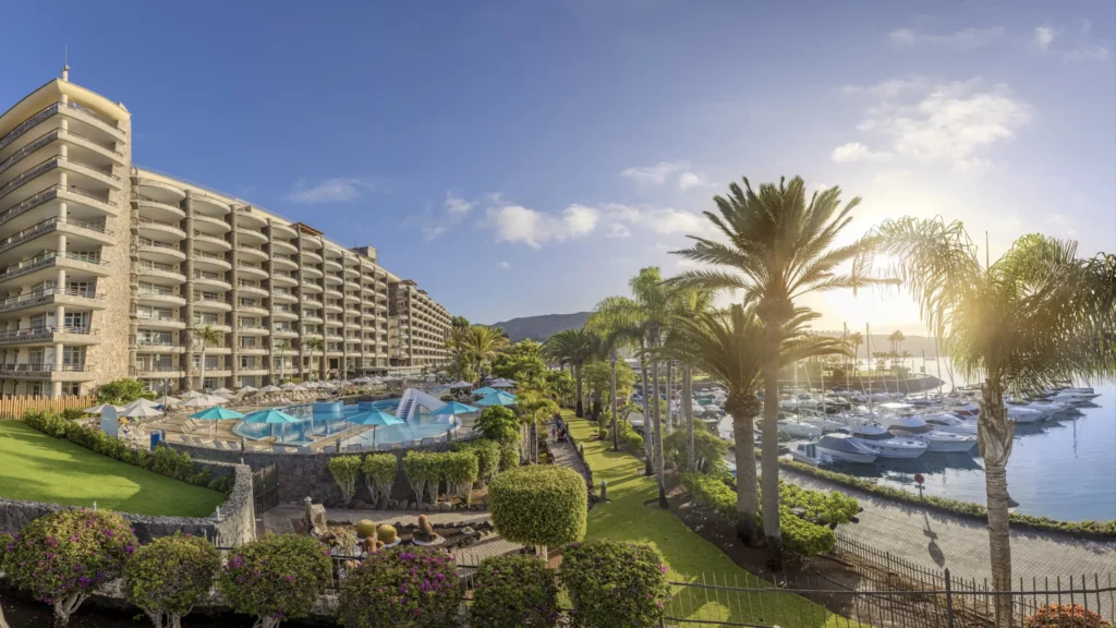 Hotel Anfi del Mar con piscina y jardines tropicales frente al puerto deportivo de Puerto de Mogán, Gran Canaria, al amanecer, con palmeras, yates y cielo despejado.
