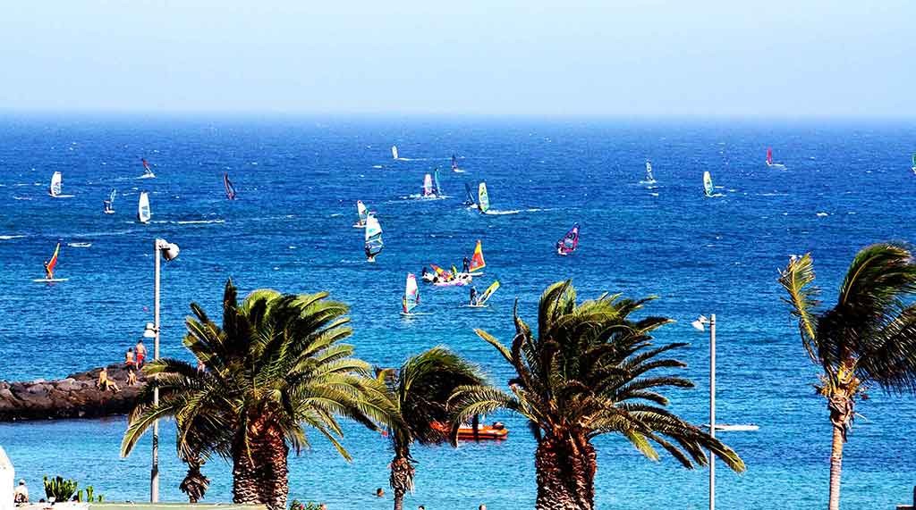 Windsurf en Playa Las Cucharas Lanzarote con velas sobre el Atlántico y el paseo marítimo al fondo