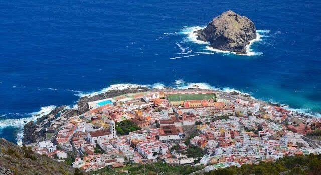 Vista aérea del pueblo de Garachico en Tenerife, Islas Canarias, mostrando edificios blancos con techos naranjas a lo largo de la costa rocosa y la distintiva formación rocosa Roque de Garachico en el profundo océano azul.